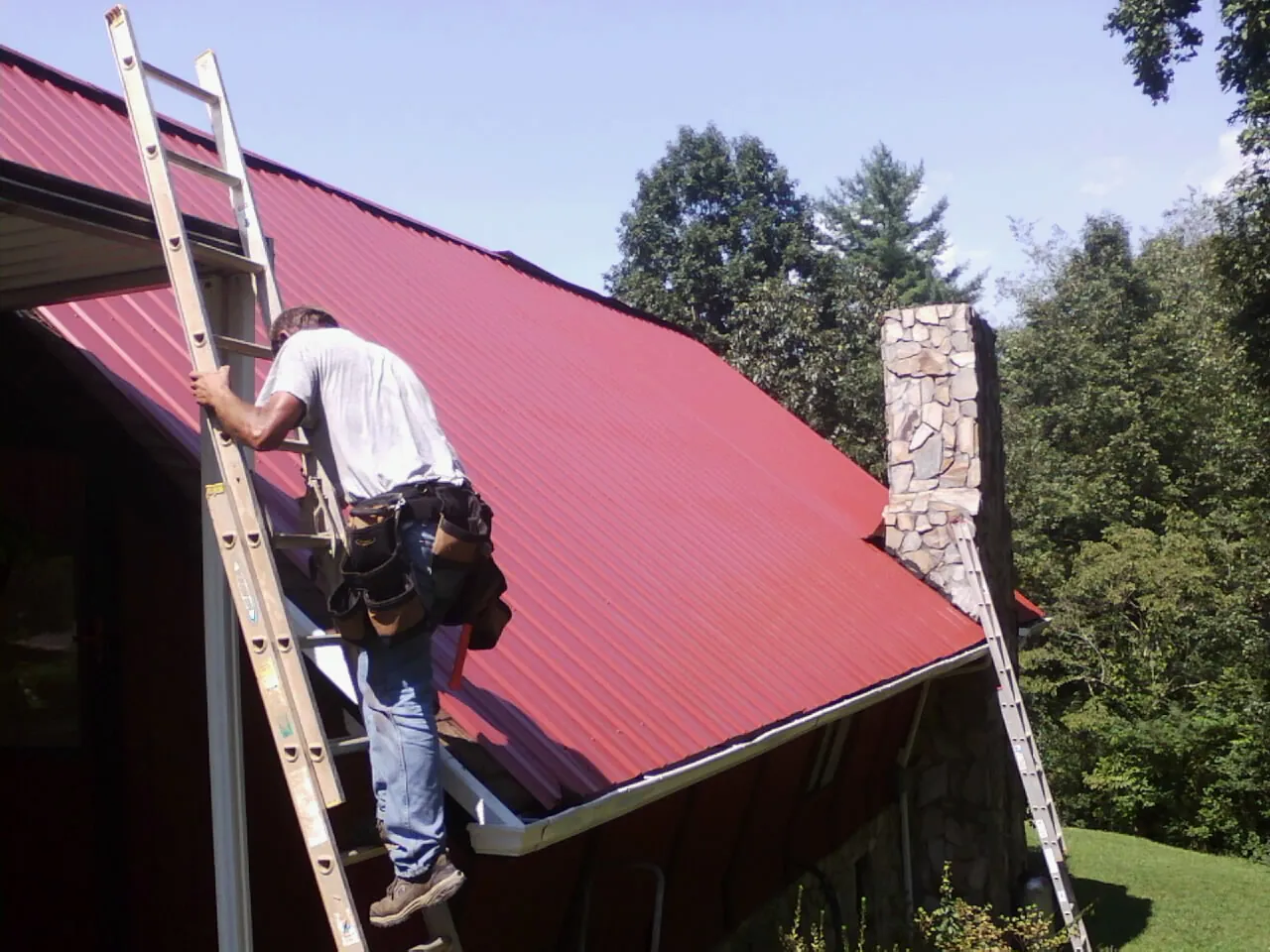 Roofer on a ladder beside a red metal roof and stone chimney — Soffit & Fascia Repair by Roofing Brothers in New Berlin
